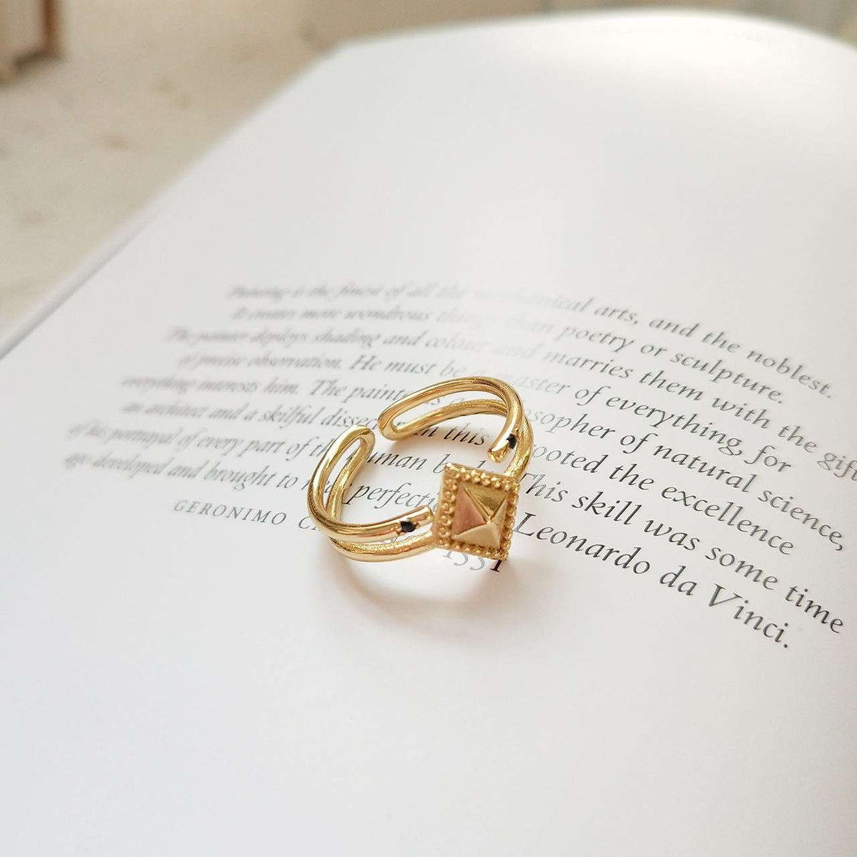 A gold ring with a triangular gold top, resting on an open book with printed text.