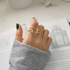 A close-up of a person's hand wearing three gold rings, including a double-helix ring and a heart-shaped ring, styled over an open book with a striped shirt cuff.