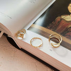 Three gold rings displayed on the pages of an open book. The rings include a square-cut stone ring, a twisted band, and a multi-loop open-style ring.