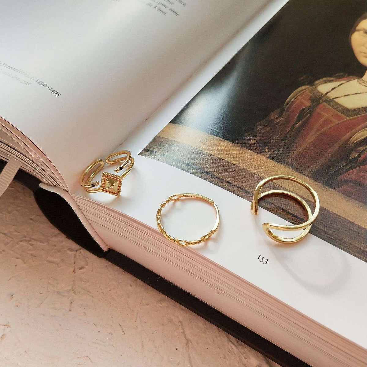 Three gold rings displayed on the pages of an open book. The rings include a square-cut stone ring, a twisted band, and a multi-loop open-style ring.