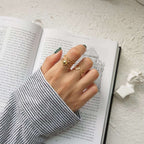 A hand wearing three gold rings, including a double-helix ring and a ring with a small red heart, resting on an open book. The person is wearing a black and white striped long-sleeve shirt, and a small white bust is visible on the textured white surface.