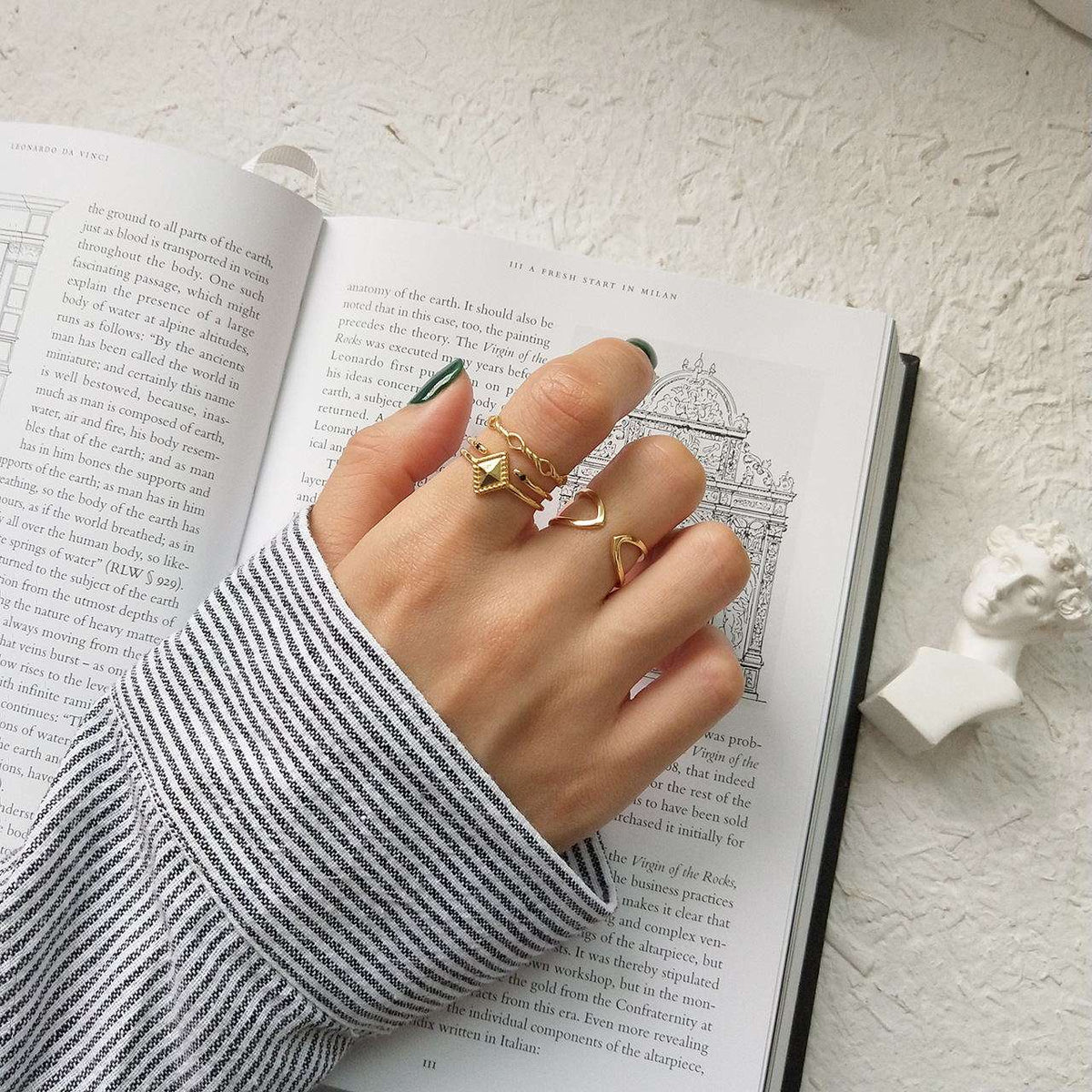 A hand wearing three gold rings, including a double-helix ring and a ring with a small red heart, resting on an open book. The person is wearing a black and white striped long-sleeve shirt, and a small white bust is visible on the textured white surface.