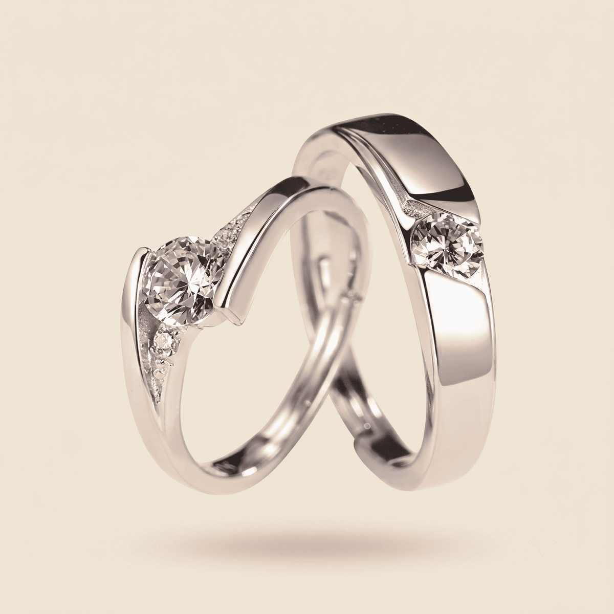 Two polished silver wedding bands with round-cut clear gemstones, shown interlocking on a warm beige background.