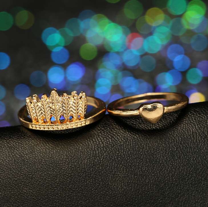 Two gold rings displayed on a black textured surface with colorful bokeh lights in the background. One ring features a crown design with small clear stones, and the other has a polished heart-shaped stone on a simple band.