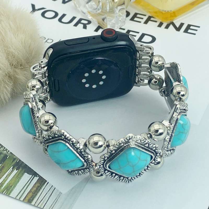 A silver-toned watch band featuring rectangular turquoise stones and polished silver beads, displayed on a watch with the back facing the camera. The watch is styled on a white surface with decorative props including a fluffy cream pom-pom and printed paper.