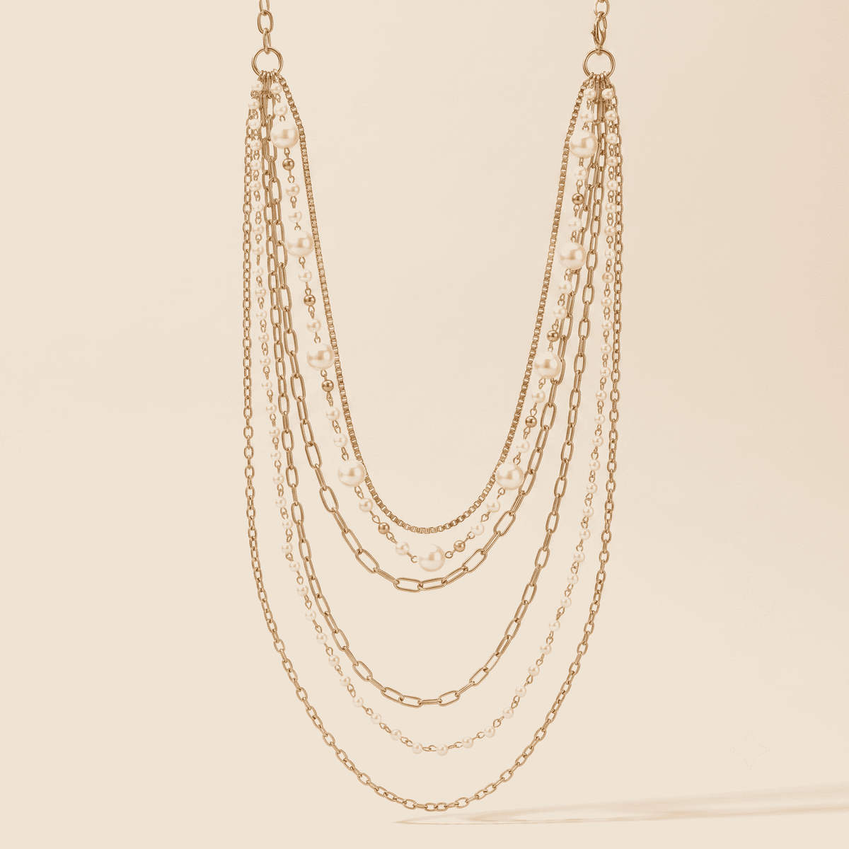 A multi-strand gold-tone chain necklace with layered paperclip, cable, and delicate chain details, displayed against a solid cream background.