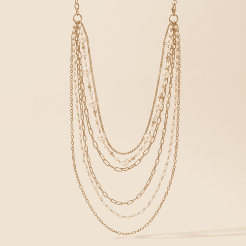 A multi-strand gold-tone chain necklace with layered paperclip, cable, and delicate chain details, displayed against a solid cream background.