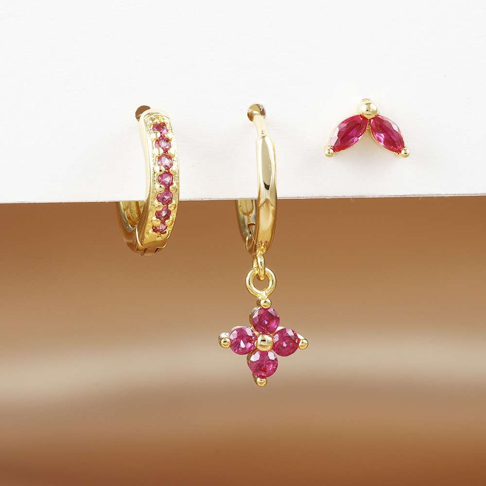 A pair of gold hoop earrings with pink gemstone accents, shown on a white display stand against a warm brown gradient background. One earring is a huggie hoop with small pink stones along the band, and the other is a dangle with a gold hoop and a dangling flower-shaped cluster of pink gemstones.