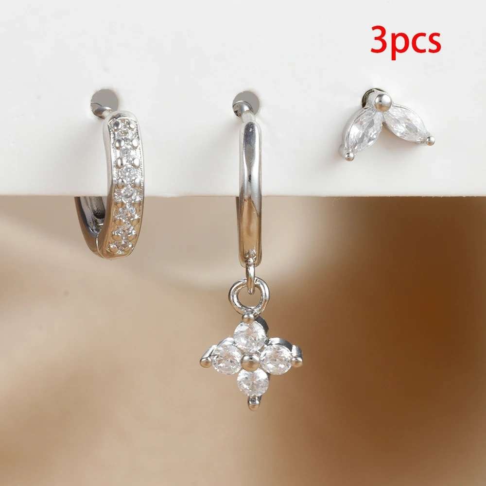 Three-piece silver stud and hoop earring set featuring a huggie hoop with clear crystals, a single drop hoop with a flower-shaped crystal pendant, and a small wing-shaped crystal stud, displayed on a white stand with a beige background.