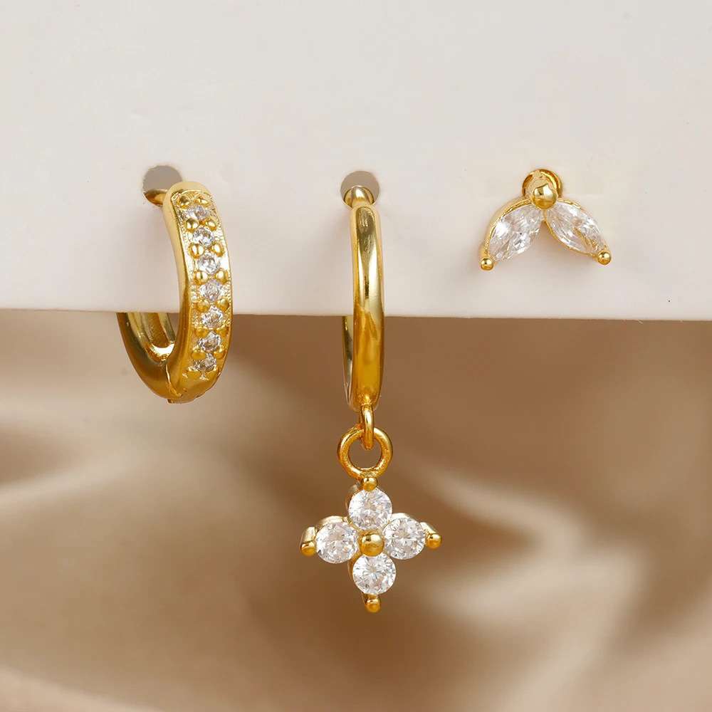 Gold hoop earrings with clear crystal accents, including one huggie hoop with a row of crystals, one hoop with a dangling flower-shaped crystal cluster, and a small matching crystal stud, displayed on a white surface with a beige satin background.