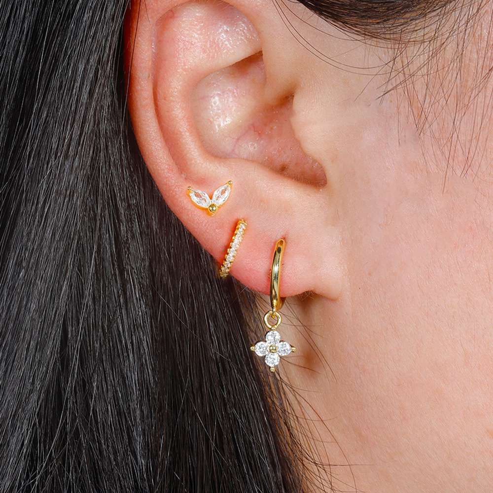 A close-up of a person's ear showcasing a stack of gold hoop and dangle earrings with clear stones, including a small butterfly earring, a huggie hoop, a curved pavé hoop, and a dangling flower-shaped earring.