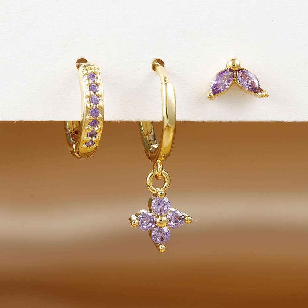 Three pairs of gold and purple gemstone earrings displayed on a white stand against a warm brown gradient background. The set includes a small huggie hoop with purple stones, a larger huggie hoop with a dangling flower-shaped drop, and a matching pair of small leaf-shaped drop earrings.