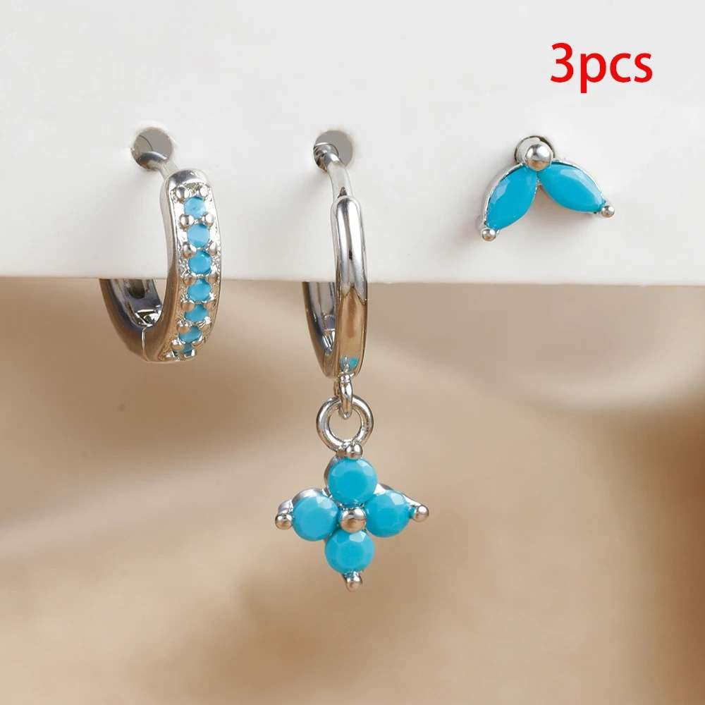 Three pairs of silver hoop earrings with bright turquoise blue stones, including one huggie hoop with a row of blue stones, one hoop with a dangling blue flower charm, and one pair of blue teardrop studs, shown on a beige background with a red '3pcs' text overlay.