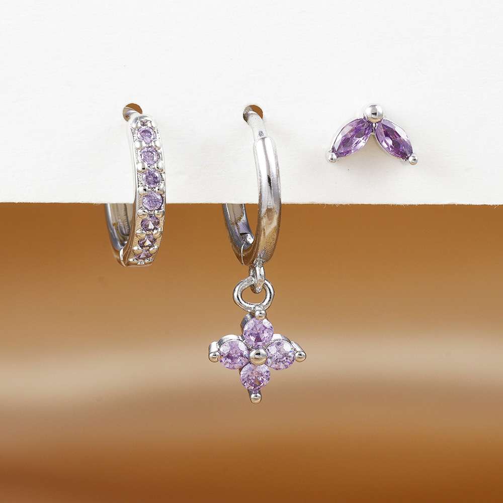 Silver hoop earrings with light purple gemstone accents, including one huggie hoop with a curved row of small purple stones, one hoop with a dangling flower-shaped purple gemstone charm, and a matching stud earring, displayed on a white surface over a warm brown gradient background.
