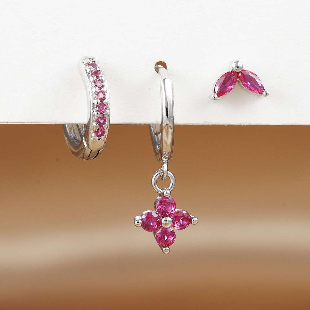 A pair of silver hoop earrings with small pink gemstones, shown alongside matching stud earrings on a white display stand against a warm brown gradient background.