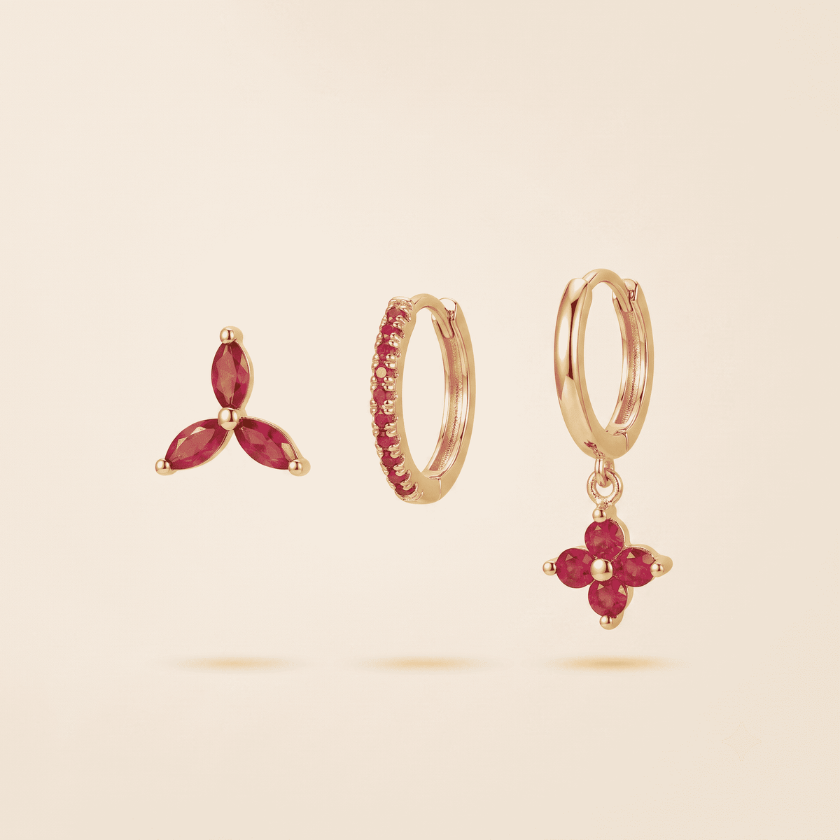 Three gold earrings with pink gemstones displayed on a light beige background: a small three-stone stud, a huggie hoop with a row of pink stones, and a hoop with a dangling flower-shaped drop set with pink stones.