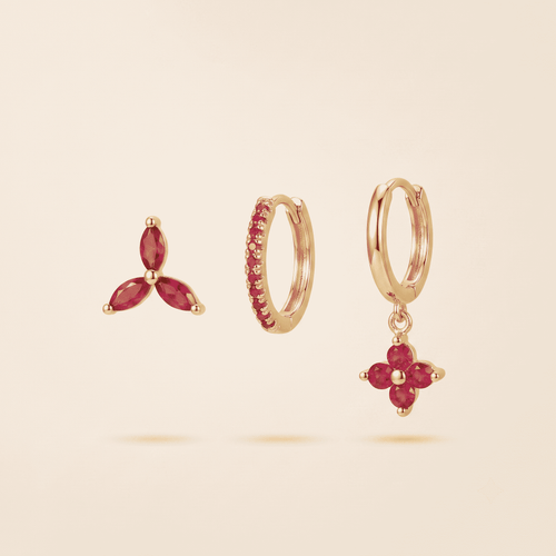 Three gold earrings with pink gemstones displayed on a light beige background: a small three-stone stud, a huggie hoop with a row of pink stones, and a hoop with a dangling flower-shaped drop set with pink stones.