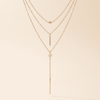 A delicate three-layer gold necklace with a long dangle bar, a small heart, and a tiny bead accent, displayed against a solid cream background.