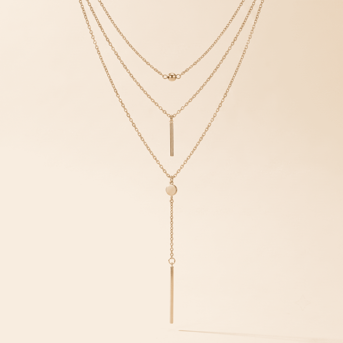 A delicate three-layer gold necklace with a long dangle bar, a small heart, and a tiny bead accent, displayed against a solid cream background.