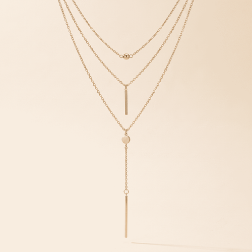 A delicate three-layer gold necklace with a long dangle bar, a small heart, and a tiny bead accent, displayed against a solid cream background.