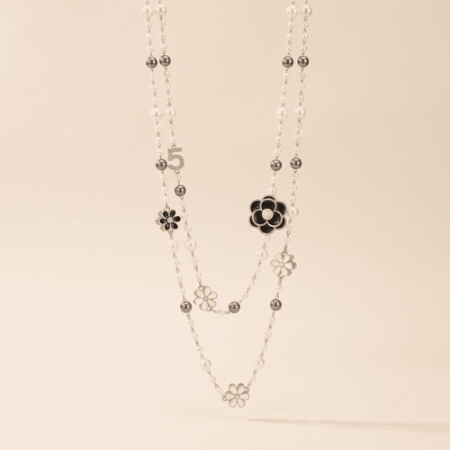 A double-strand silver necklace featuring white pearl-like beads, small silver flower charms, and one larger black-and-white flower accent, displayed against a solid light beige background.