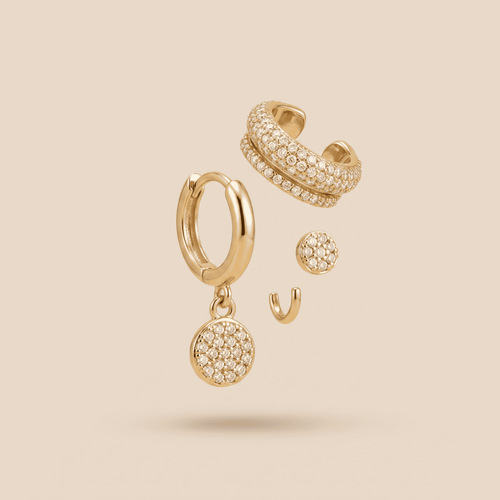 Gold-toned huggie hoop earring with a round pavé disc drop, shown alongside matching stud and stacked pavé hoop earrings on a warm beige background.