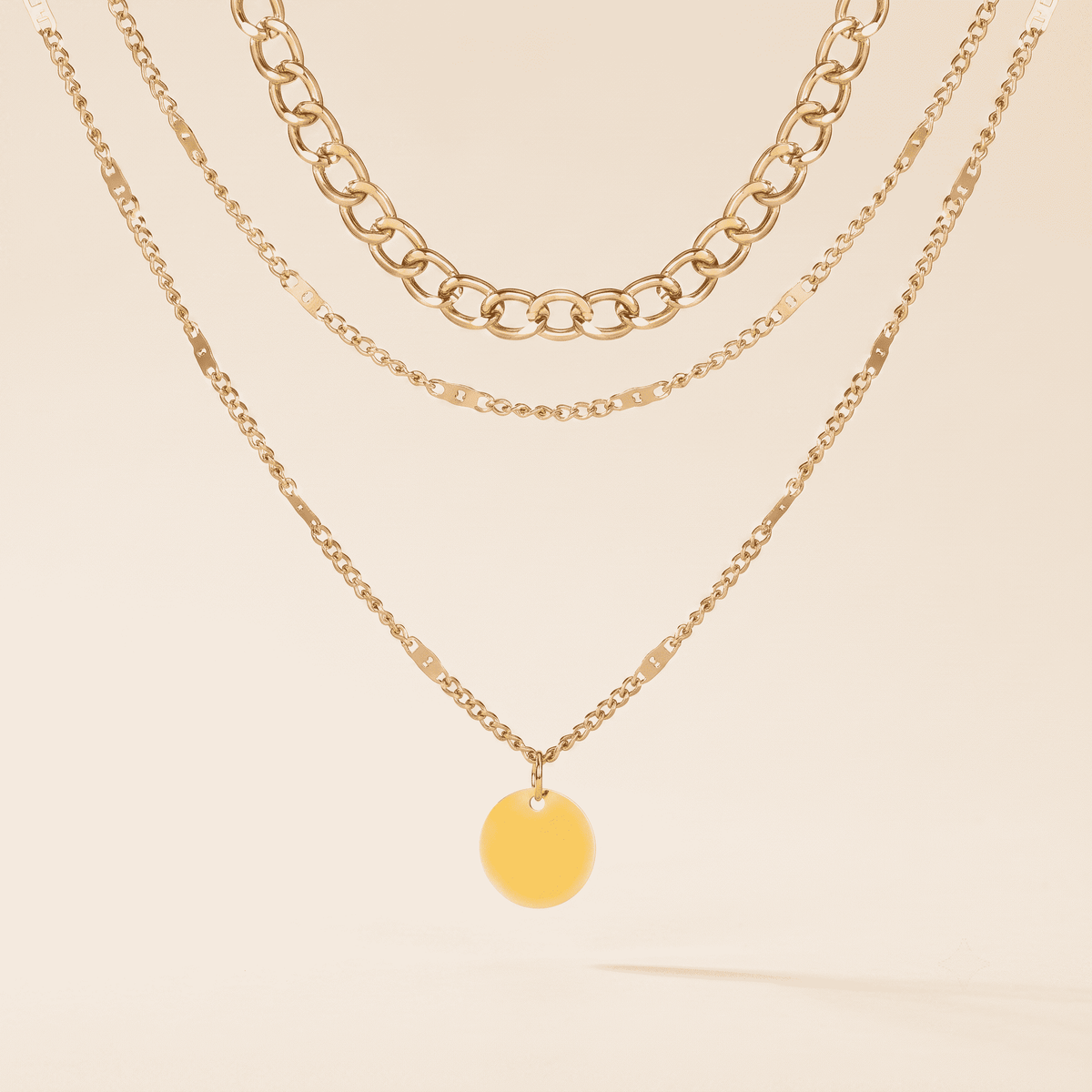 Three layered gold-tone necklaces displayed on a warm cream background: a chunky curb chain, a fine link chain, and a delicate chain with a round yellow-gold disc pendant.