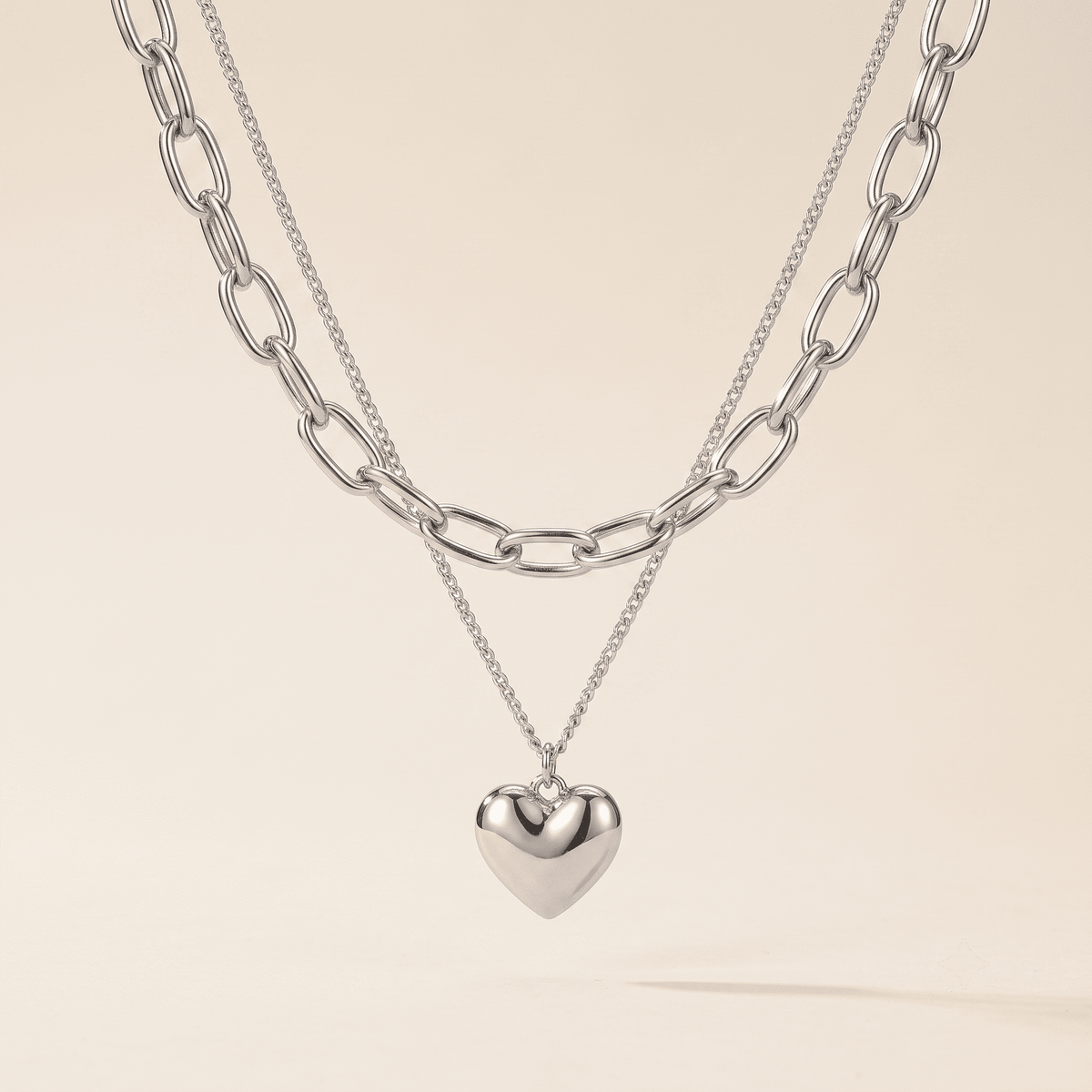 A layered silver necklace set featuring a chunky paperclip chain, a delicate chain with a polished heart pendant, and a small heart charm, displayed against a solid light beige background.