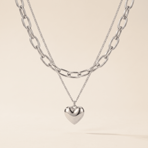 A layered silver necklace set featuring a chunky paperclip chain, a delicate chain with a polished heart pendant, and a small heart charm, displayed against a solid light beige background.