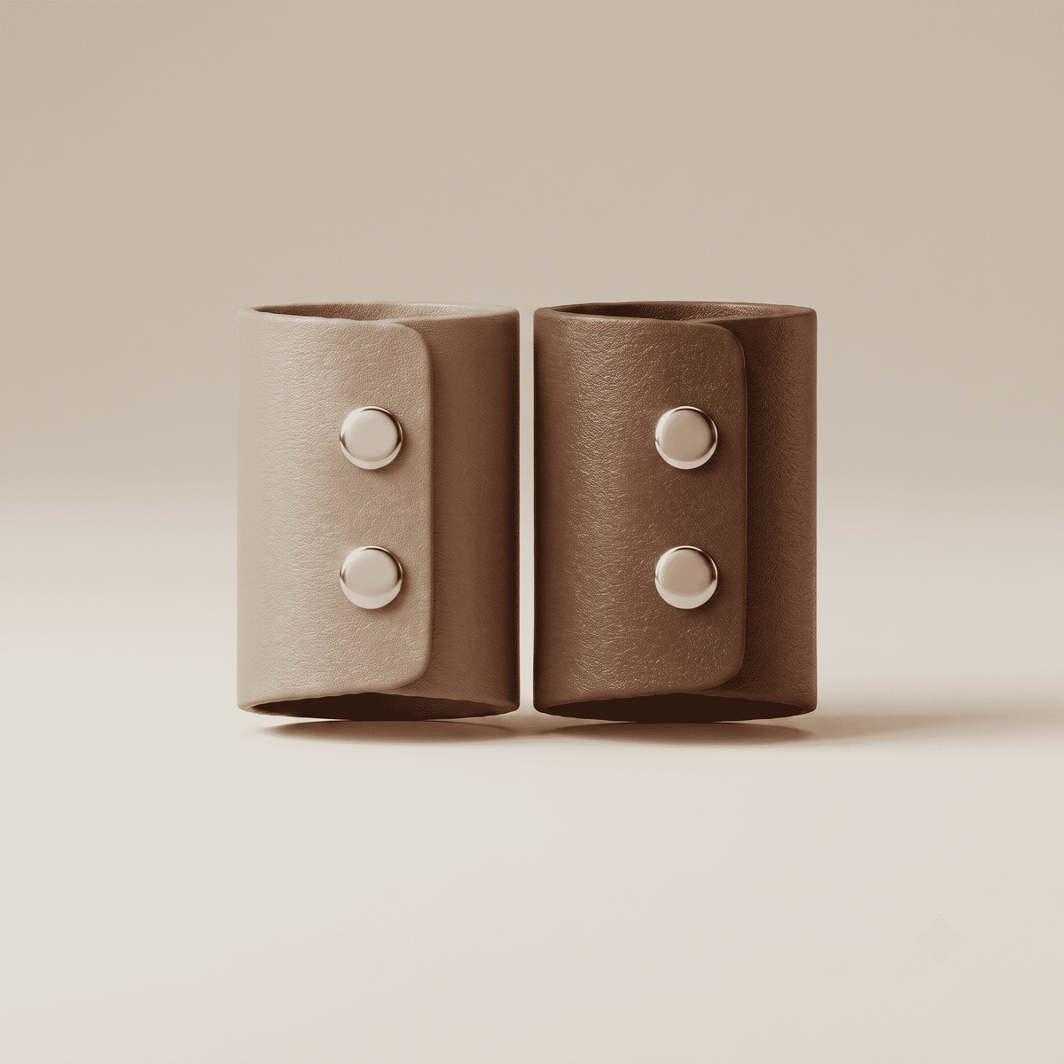 Two leather cuff bracelets in taupe and brown tones, each featuring two silver-toned snap buttons, displayed side-by-side on a warm beige background.