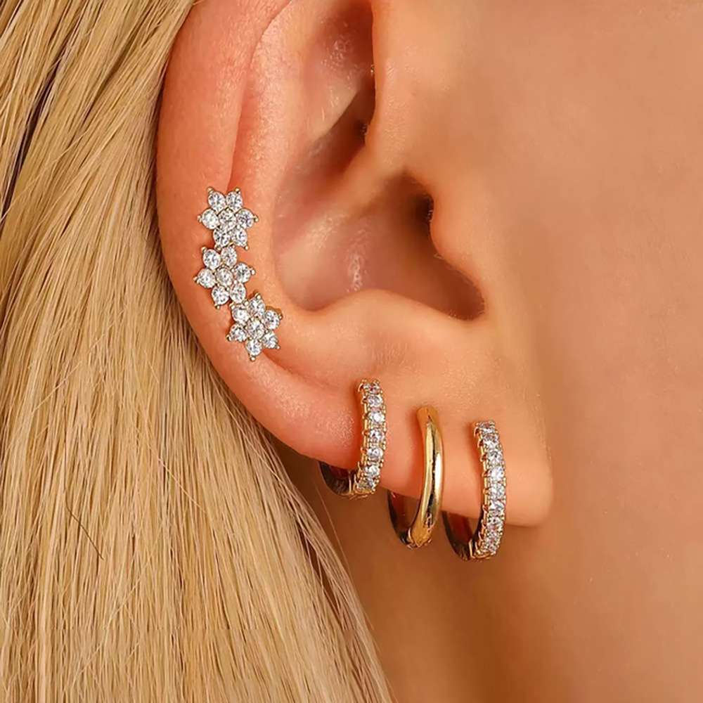 A close-up of a person's ear wearing multiple earrings: a silver-toned ear cuff with three flower clusters, a small silver huggie hoop, a gold huggie hoop, and a silver huggie hoop set with clear stones.