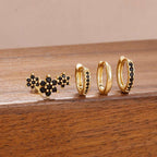 Three pairs of gold hoop and stud earrings with small black stones, displayed on a wooden surface against a beige background.