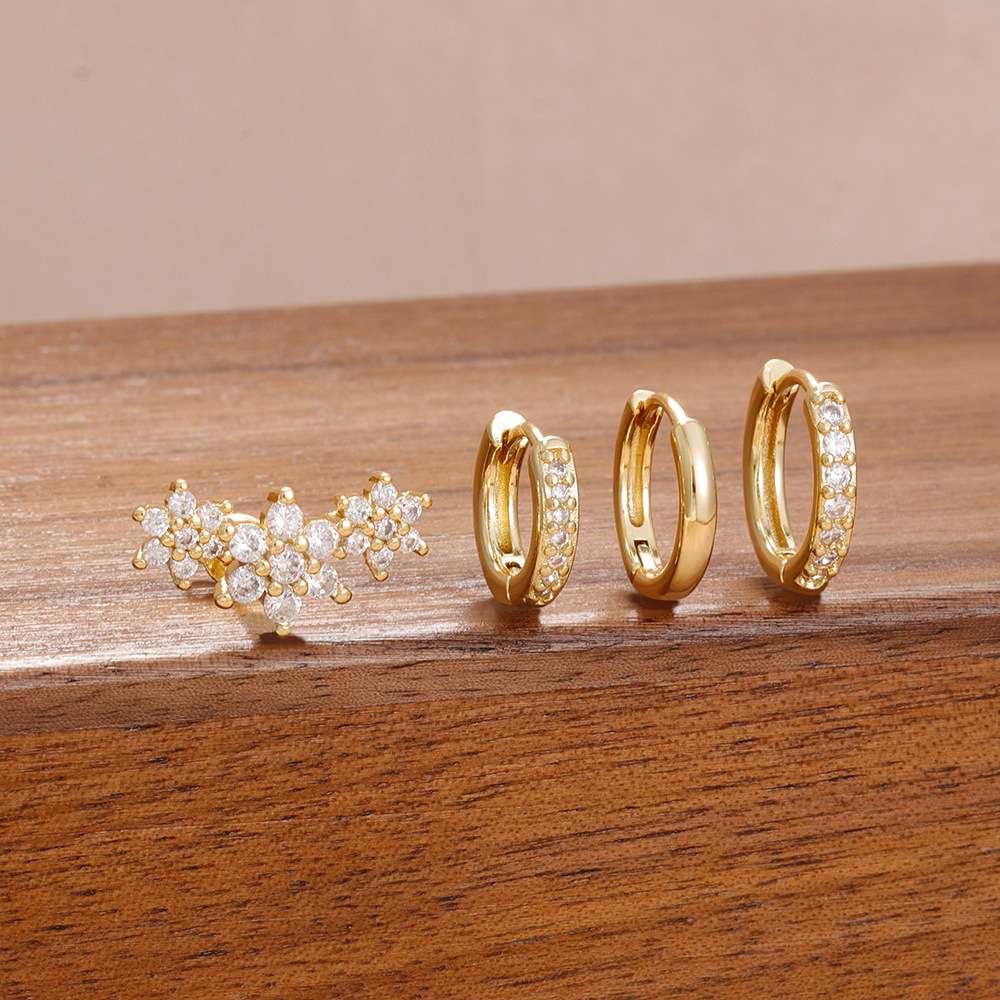 Three pairs of gold-toned jewelry displayed on a wooden surface: a floral cluster stud earring, a small hoop earring, and a huggie hoop earring with clear stones along the front.
