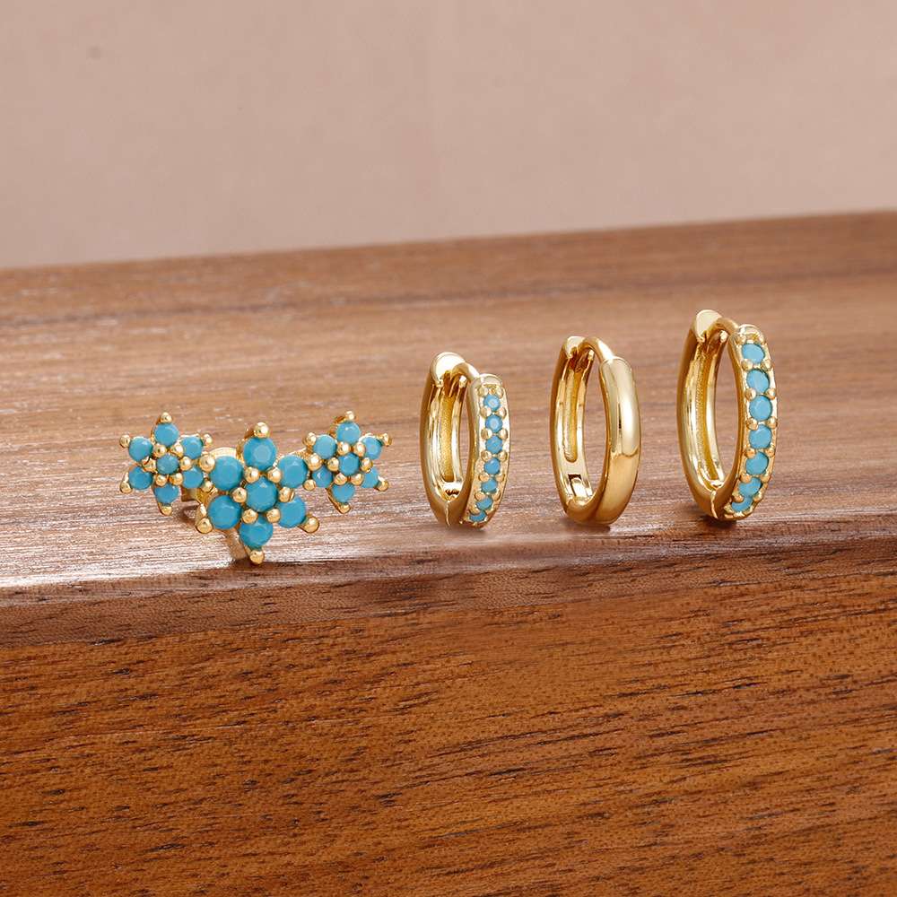 A set of gold jewelry displayed on a wooden surface, including a cluster stud earring with turquoise blue stones and two huggie hoop earrings with matching blue stone accents.