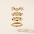 A set of gold rings with clear gemstones, including a cluster of starburst rings at the top, two pave-set hoop rings in the middle, and one smooth band ring at the bottom, displayed on a light beige background.