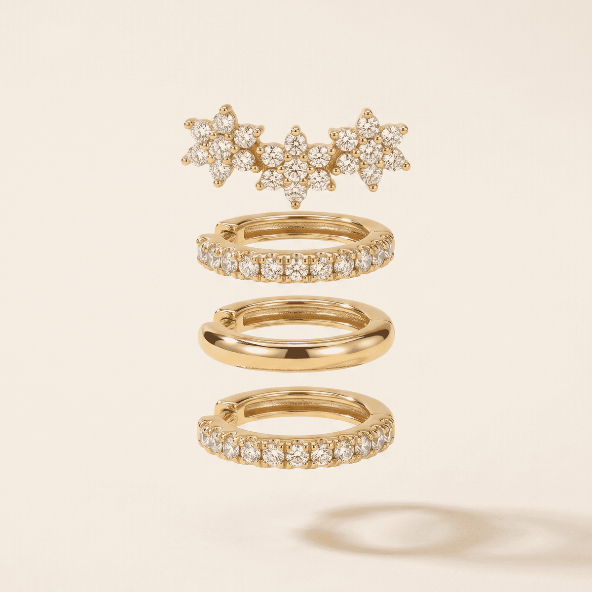 A set of gold rings with clear gemstones, including a cluster of starburst rings at the top, two pave-set hoop rings in the middle, and one smooth band ring at the bottom, displayed on a light beige background.