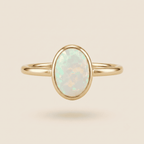 A gold ring with an oval-shaped white opal gemstone, shown against a solid light beige background.