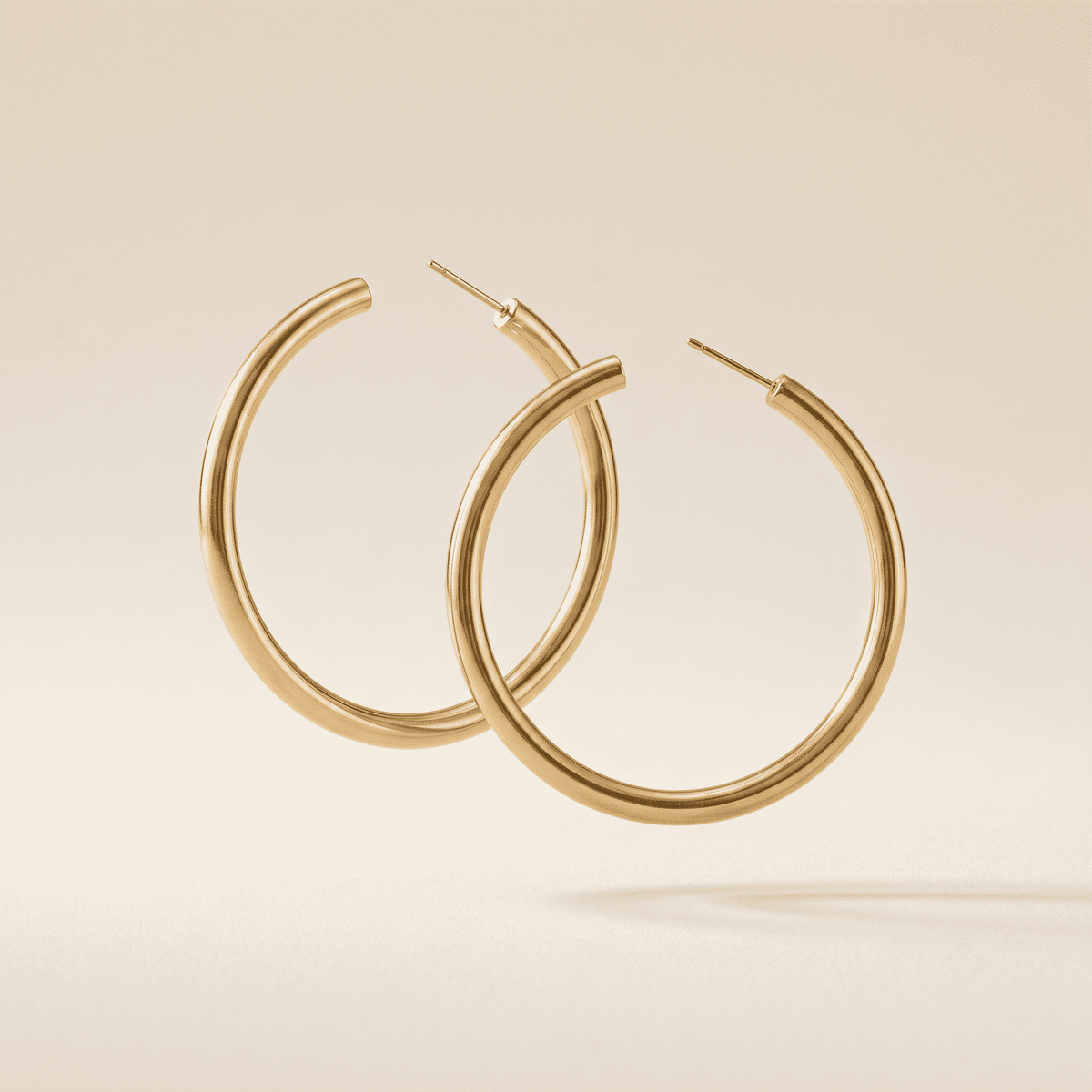 A pair of thick, polished gold hoop earrings floating against a warm beige background.