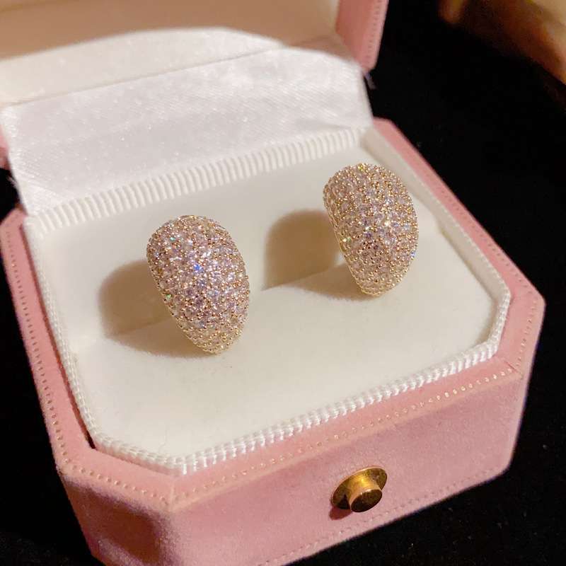 A pair of gold-toned dome stud earrings covered in sparkling clear stones, displayed inside an open pink jewelry box with a white velvet interior.