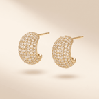 A pair of gold crescent-shaped hoop earrings paved with small clear stones, shown on a warm beige background.