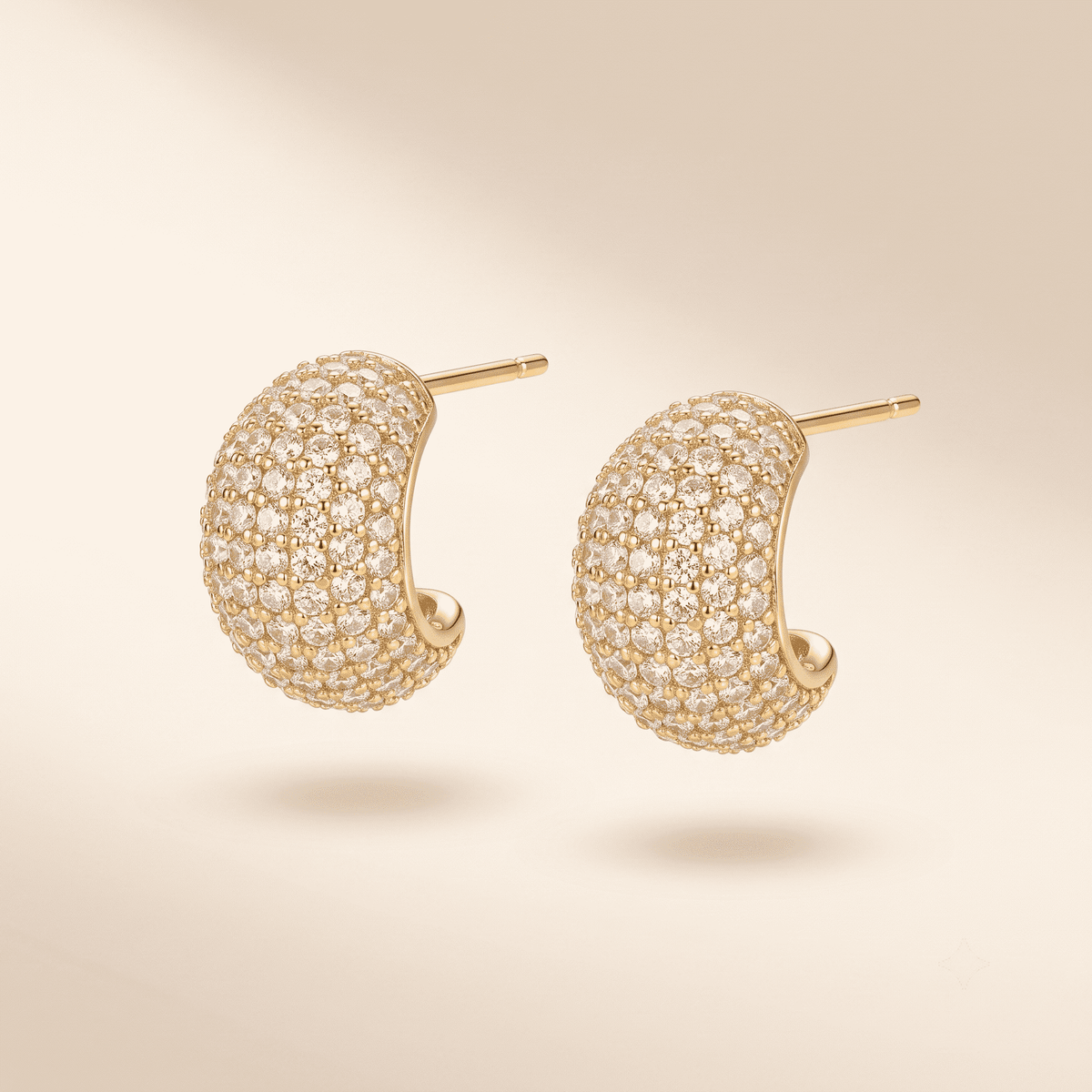 A pair of gold crescent-shaped hoop earrings paved with small clear stones, shown on a warm beige background.