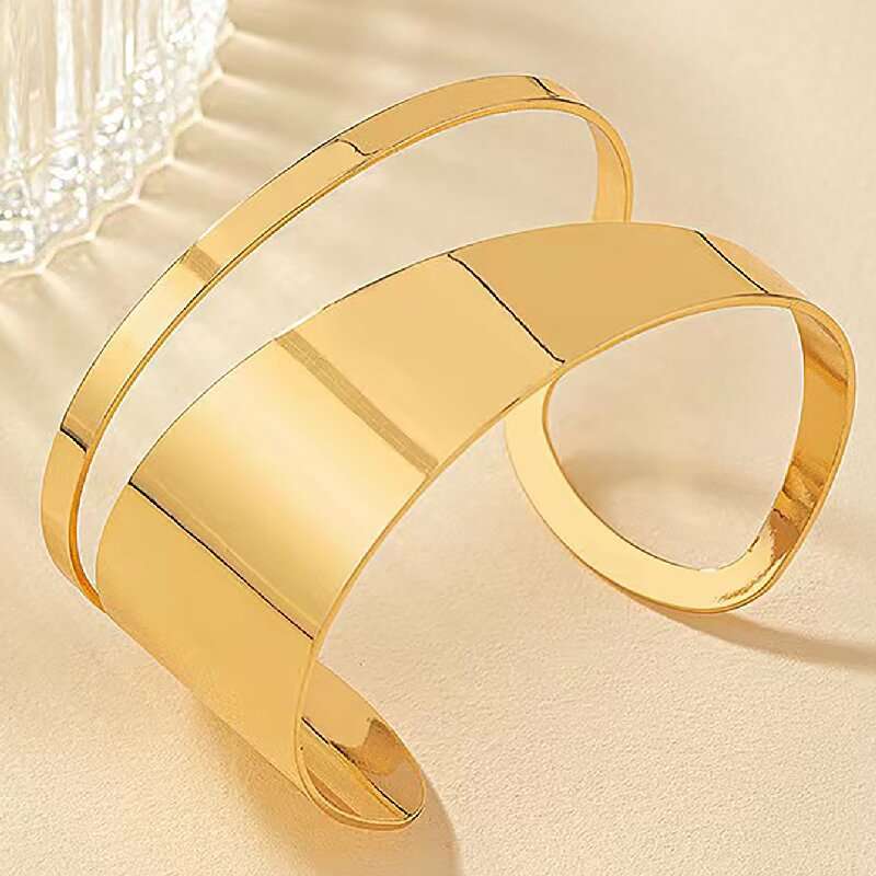 A polished gold-tone metal cuff bracelet with a modern, layered open-band design, displayed on a textured beige surface.