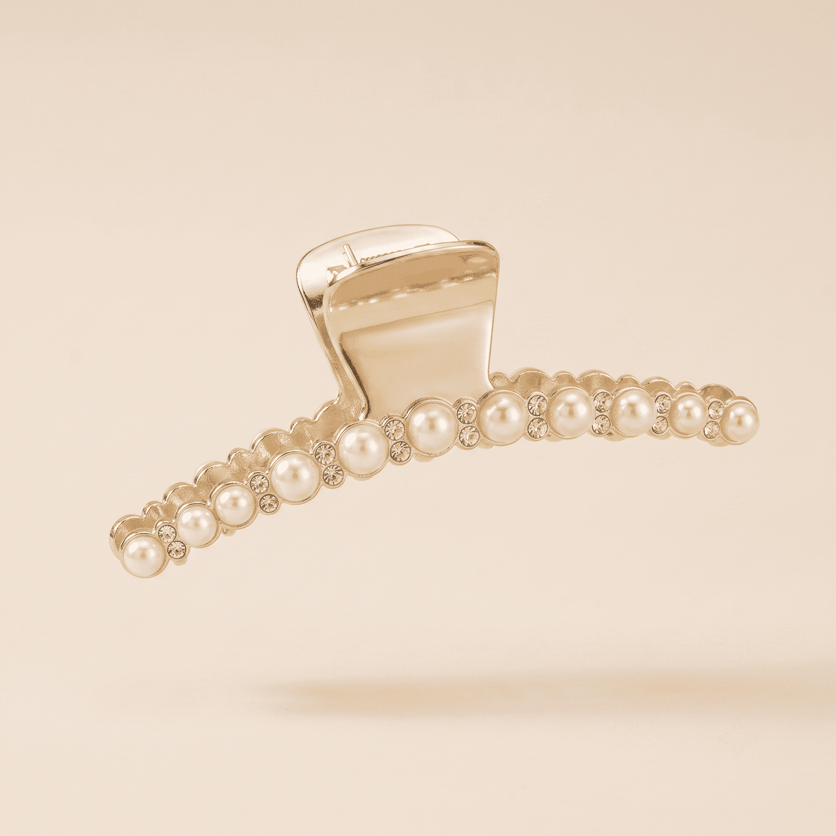 A gold-toned hair claw clip embellished with small white pearls and clear rhinestones, shown on a solid beige background.