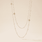 A double-strand pearl necklace with gold rose and geometric charms, displayed against a warm beige background.