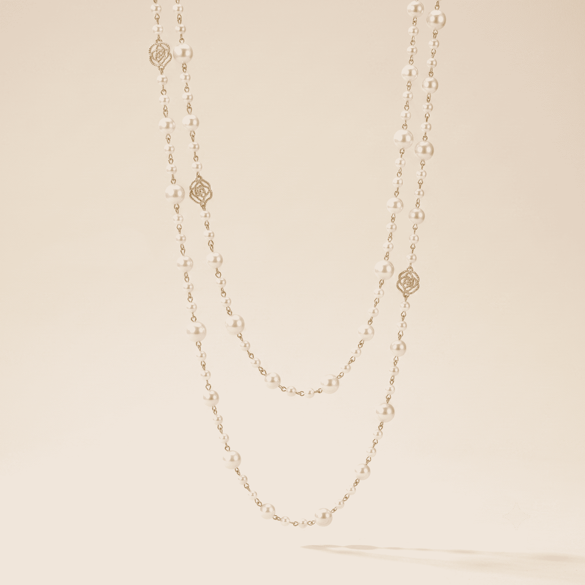 A double-strand pearl necklace with gold rose and geometric charms, displayed against a warm beige background.
