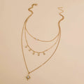 A gold-toned multi-layered necklace featuring three delicate chains, star-shaped charms, and a larger starburst pendant with a clear rhinestone, displayed on a solid beige background.
