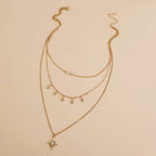 A gold-toned multi-layered necklace featuring three delicate chains, star-shaped charms, and a larger starburst pendant with a clear rhinestone, displayed on a solid beige background.