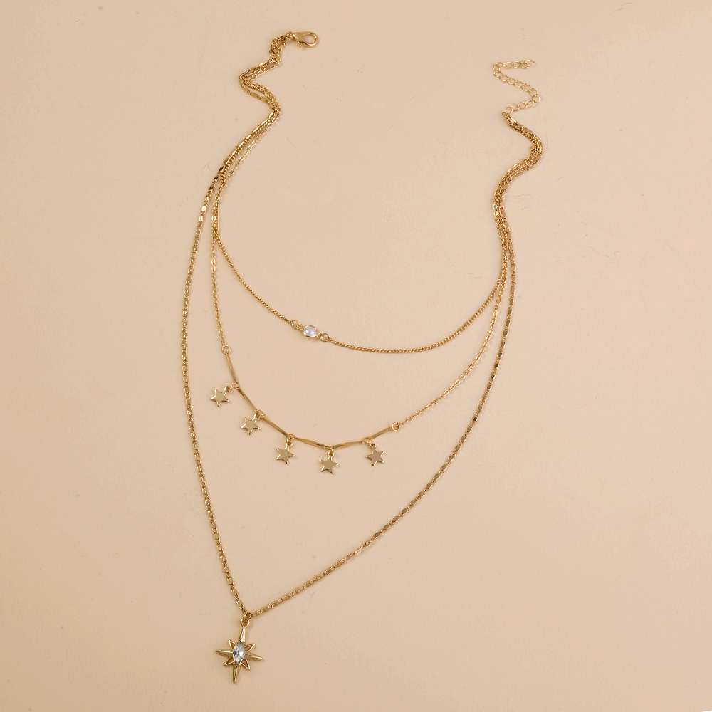 A gold-toned multi-layered necklace featuring three delicate chains, star-shaped charms, and a larger starburst pendant with a clear rhinestone, displayed on a solid beige background.