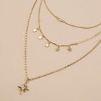 A layered gold necklace set featuring three delicate chains, including star-shaped pendants, a beaded chain, and a larger chain with a starburst charm set with a clear crystal, displayed on a solid beige background.
