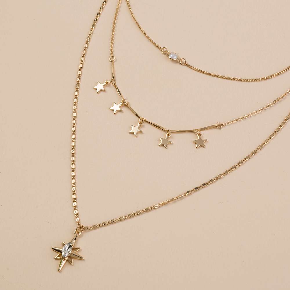 A layered gold necklace set featuring three delicate chains, including star-shaped pendants, a beaded chain, and a larger chain with a starburst charm set with a clear crystal, displayed on a solid beige background.
